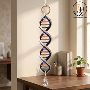 May include: A crocheted DNA double helix wall hanging suspended from a light wood ring. The structure features cream, navy blue, and multi-colored yarn segments. A clear, faceted teardrop-shaped crystal hangs from the bottom of the fiber art piece.