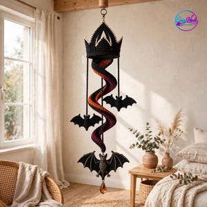 May include: A hanging crochet mobile featuring a black crown at the top, a central spiral transitioning from orange to dark purple, and three black crocheted bats attached at different levels. A small amber-colored crystal hangs at the bottom.