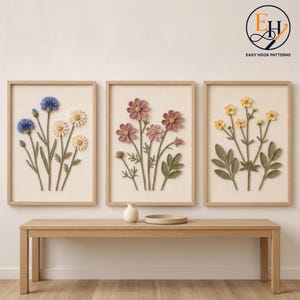 May include: Three rectangular framed wall art pieces featuring textured, three-dimensional floral arrangements on a cream background. The left frame shows blue and white flowers, the center frame displays pink flowers, and the right frame shows yellow flowers.