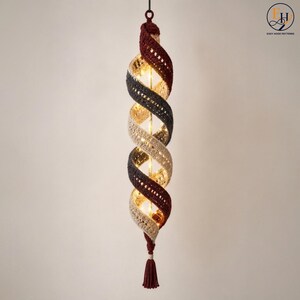 Peut inclure: Une décoration suspendue en spirale avec une texture crochetée en bandes alternées de rouge foncé, gris anthracite et crème. La structure est éclairée de l'intérieur par une guirlande verticale de lumières blanc chaud et terminée par un pompon.