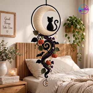 May include: A crochet wall hanging featuring a large cream-colored crescent moon with a black cat silhouette sitting on top. A black hoop frame holds the moon, with dangling crocheted vines, orange pumpkins, small purple berries, and black bats.
