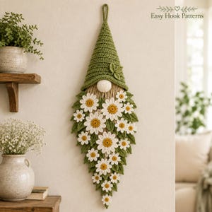 Op de afbeelding: Een wandhanger met een gehaakte kabouter met een hoge groene muts en een baard gemaakt van witte madeliefjes met gele harten en groene bladeren. De kabouter heeft een kleine ronde neus en een lus aan de bovenkant om op te hangen. Tekst: Easy Hook Patterns.