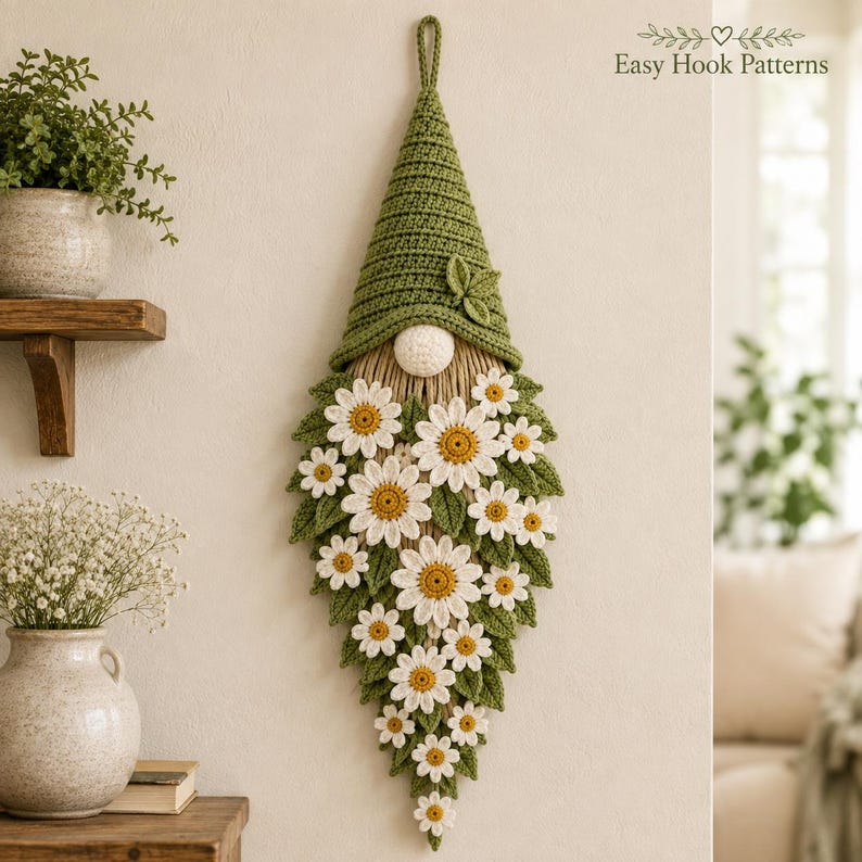 Op de afbeelding: Een wandhanger met een gehaakte kabouter met een hoge groene muts en een baard gemaakt van witte madeliefjes met gele harten en groene bladeren. De kabouter heeft een kleine ronde neus en een lus aan de bovenkant om op te hangen. Tekst: Easy Hook Patterns.