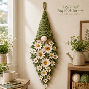 Op de afbeelding: Een gehaakte wandhanger in de vorm van een kabouter met een hoge olijfgroene muts, een ronde witte neus en een lange baard versierd met talloze witte en gele madeliefjes en groene bladeren. De tekst Easy Hook Patterns is duidelijk zichtbaar.