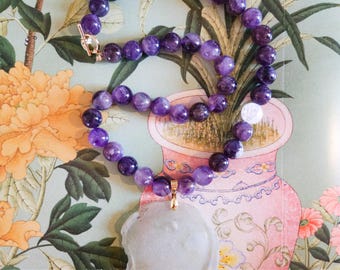 May include: A necklace featuring a strand of round, deep purple amethyst beads and a single white bead, connected to a large, circular, pale grey pendant with a smooth, irregular edge. The pendant is attached with a gold-toned metal bail.