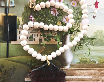 May include: A necklace featuring a strand of smooth, round, light-colored wooden beads with a single pale pink bead and a large, dark, irregular-shaped pendant. The necklace is secured with a gold-toned toggle clasp at the top.