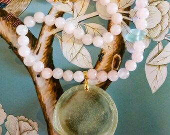 May include: A necklace featuring a strand of pale pink spherical beads and a large, circular, translucent light green pendant. The pendant is attached to the bead strand by a gold-toned metal bail, and the necklace includes a gold-toned clasp.