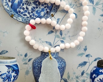 May include: A necklace featuring a strand of pale pink spherical beads with a single bright red bead accent. A large, translucent, irregular white pendant hangs from a gold-toned metal bail at the center of the beaded strand.