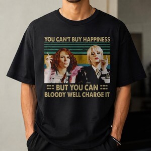 May include: A black short-sleeved t-shirt featuring a graphic print of two individuals with the text "YOU CAN'T BUY HAPPINESS BUT YOU CAN BLOODY WELL CHARGE IT" set against a background of horizontal stripes in shades of green, yellow, and blue.