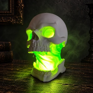 Op de afbeelding: Een witte schedelvormige lamp met een uitgeholde schedel en kaak die van binnenuit een levendig, gloeiend groen licht uitstraalt. De schedelstructuur is matwit en contrasteert met de heldere neongroene verlichting die uit de oogkassen en de nekregio komt.