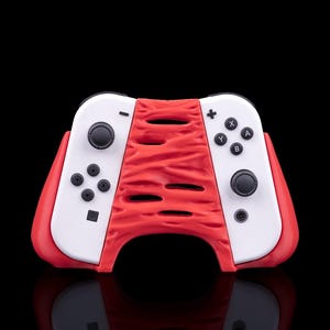 Pode incluir: Um comando de videojogos branco com uma pega central vermelha, texturada e de forma orgânica com recortes horizontais. O comando inclui joysticks pretos, um pad direcional e botões com os símbolos X, Y, A e B no lado direito.