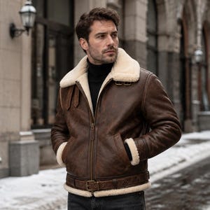 May include: A brown leather bomber jacket with a thick cream-colored shearling collar, cuffs, and hem. The jacket features a front zipper closure, two side pockets, and a buckled belt at the waist, worn over a black turtleneck sweater.