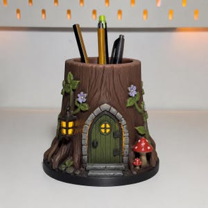May include: A brown tree stump pencil holder featuring a green arched door with a small window, a hanging yellow lantern, purple flowers on vines, and two red mushrooms with white spots. Three pens are inserted into the top opening of the holder.
