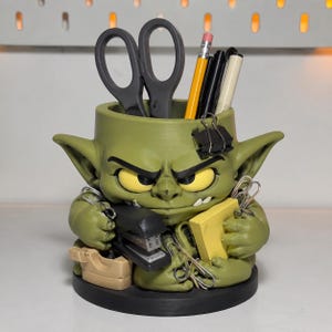 May include: A green desk organizer shaped like a grumpy creature with large yellow eyes and pointed ears. The figure holds a black stapler and a yellow notepad, while the top opening contains black scissors, a yellow pencil, and pens.
