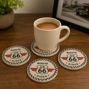 May include: A set of four round ceramic coasters featuring a Route 66 US Diner graphic with a black and white checkered border, red wings, and red stars. One coaster supports a plain white ceramic mug filled with a light brown beverage.