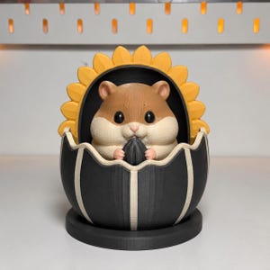 Pode incluir: Uma estatueta impressa em 3D de um hamster castanho e creme sentado dentro de uma casca em forma de ovo preta e branca. O hamster segura uma semente de girassol preta nas patas. Atrás do hamster há uma moldura de pétalas de girassol amarelas.