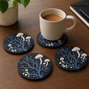 May include: Three circular black coasters featuring a raised blue tree branch design with white mushrooms. One coaster supports a beige ceramic mug filled with coffee. The coasters have a matte finish and a detailed, textured, relief-style surface.
