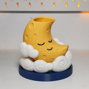 May include: A yellow crescent moon planter with a sleeping face, featuring crater-like indentations on its surface. The moon rests within a base of white, swirled clouds, all supported by a solid, circular dark blue stand for stability.