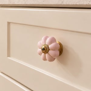 May include: A set of light pink, pumpkin-shaped ceramic cabinet knobs with a brass-toned metal centre and base. These decorative knobs are installed on cream-coloured kitchen cabinets, featuring a fluted, rounded design with a glossy finish.