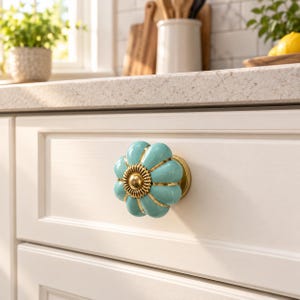 May include: A turquoise ceramic cabinet knob with a scalloped, flower-like shape and a central gold-toned metal accent. The knob features a glossy finish and is mounted on a white drawer front, showcasing a metallic gold base and trim.