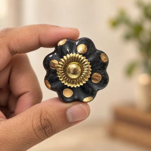 May include: A black ceramic cabinet knob with a scalloped, flower-like shape. The surface features gold-coloured polka dots and a central, circular gold-toned metal accent with a ridged, sunburst-style edge and a smooth, raised centre dome.