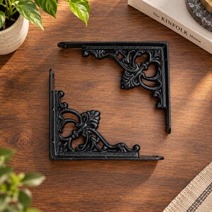 Può includere: Due reggimensola a forma di L di colore nero con un intricato e ornato design a volute. Ogni supporto presenta una superficie di montaggio piatta con fori preforati e un braccio di supporto curvo e decorativo che unisce le sezioni.