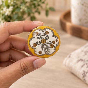 Könnte beinhalten: Ein runder Keramikknopf mit gewelltem Rand und einer leuchtend gelben Umrandung. Die Mitte ist weiß und mit einem detaillierten botanischen Muster in Dunkelgrau und Senfgelb verziert, das kleine Blumenmotive und Blattreben zeigt.