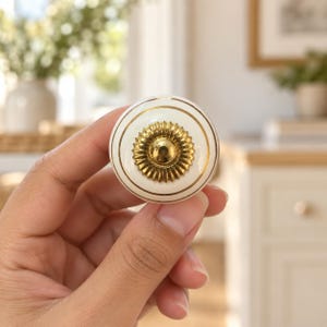 May include: A round cabinet knob featuring a smooth cream-colored surface accented with thin gold circular lines. The center of the knob is adorned with a raised, metallic gold circular medallion that has a textured, sunburst-like pattern.