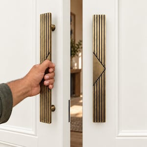 May include: Two long, vertical door handles made of brushed brass with a dark, linear, ribbed texture. Each handle features a central triangular cutout design. The handles are mounted on white double doors, with one handle being gripped by a hand.