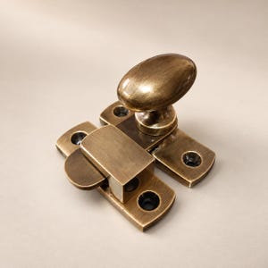 Puede incluir: Un pestillo de ventana de latón con un pomo ovalado unido a una placa base rectangular con cuatro orificios para tornillos avellanados. El herraje metálico tiene un acabado cepillado y envejecido con pátina oscura visible en la superficie.