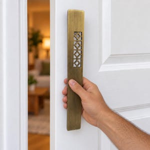 May include: A long, rectangular, brushed brass door pull handle with a vertical cutout pattern featuring five stacked diamond shapes. The metal handle is mounted vertically on a white paneled door, showcasing a metallic finish and clean lines.