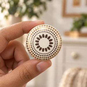 Può includere: Un pomello in ceramica rotondo con base color crema, un motivo floreale centrale marrone scuro e cerchi concentrici di punti dorati metallizzati. Il design è simmetrico, con i punti dorati che aumentano di dimensioni verso il bordo.