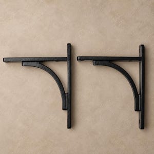 Puede incluir: Un par de soportes para estantes en forma de L hechos de metal negro. Cada soporte cuenta con una placa de montaje vertical, un brazo de soporte horizontal y un refuerzo diagonal curvo que conecta ambas secciones. El metal tiene un acabado mate texturizado.