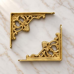 May include: Two L-shaped shelf brackets made of polished gold-toned metal. Each bracket features an intricate, ornate scrollwork design with leaf-like motifs and open cutouts. The brackets have flat mounting surfaces with visible screw holes.