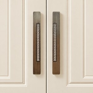 May include: Two vertical rectangular door handles made of dark bronze metal, each featuring a central recessed strip with a repeating geometric diamond and square cutout pattern, mounted on a pair of off-white panelled cabinet doors.