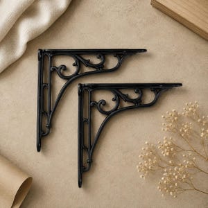 Könnte beinhalten: Zwei schwarze Regalwinkel aus Metall mit einem verzierten, geschwungenen Schnörkeldesign. Jeder Winkel verfügt über eine vertikale Montageplatte mit vorgebohrten Löchern und einen horizontalen Tragarm mit einem detaillierten Durchbruchmuster.