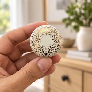 May include: A round, off-white ceramic cabinet knob featuring a metallic gold filigree pattern. The design consists of intricate, symmetrical, swirling lines arranged in a circular motif across the surface of the knob, creating a decorative accent.