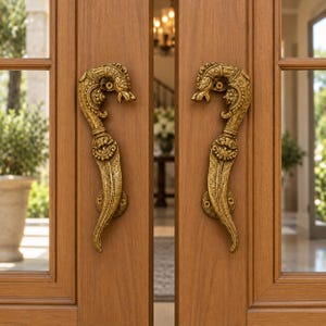 May include: A pair of ornate, gold-toned metal door handles mounted on double wooden doors. Each handle features a curved, textured design with a sculpted animal head at the top and a decorative face detail in the center, secured with visible screws.