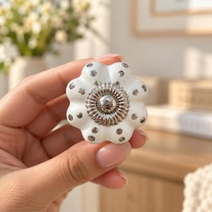 May include: A white ceramic cabinet knob shaped like a flower with six rounded petals. The surface features small, evenly spaced silver metallic dots. A circular, ridged silver metallic centre piece is fixed in the middle of the knob.