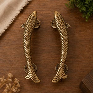 May include: A pair of brass door handles shaped like fish, featuring detailed scale textures, a curved body, and a flared tail. Each handle includes two circular mounting plates with pre-drilled screw holes at the head and tail ends for installation.