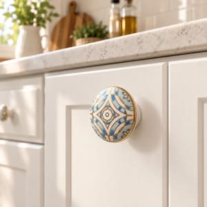 May include: A round ceramic cabinet knob featuring a symmetrical geometric pattern in shades of blue, white, and gold. The knob has a smooth, glossy finish and is mounted on a white cabinet door with a recessed panel design and gold hardware.