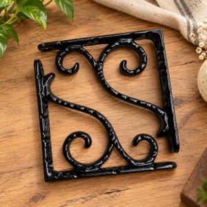 May include: A black metal shelf bracket with a textured finish, featuring a triangular frame and decorative scrollwork patterns. The bracket includes pre-drilled holes at the ends for mounting, showcasing a sturdy construction with curved metal elements.