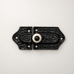 May include: A black metal latch featuring an intricate embossed pattern and a central white ceramic knob with a black dot. The hardware includes four circular screw holes for mounting, with a symmetrical design that secures two pieces together.