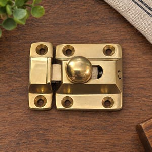 May include: A polished brass cabinet latch featuring a rectangular base plate with four screw holes and a separate strike plate with two screw holes. The mechanism includes a central spherical knob that slides horizontally to engage the latch.