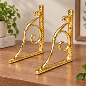 May include: Two L-shaped shelf brackets made of polished gold-tone metal. Each bracket features a vertical back plate with pre-drilled screw holes and a decorative curved support arm with a scroll design. The metal surfaces have a textured finish.