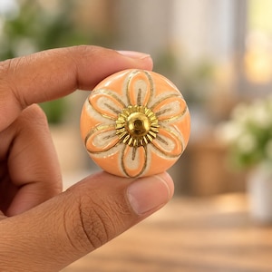 May include: A round ceramic cabinet knob featuring a peach-coloured floral pattern with cream-coloured petals outlined in gold. The centre of the flower is adorned with a small, circular, metallic gold-toned decorative cap with a scalloped edge.