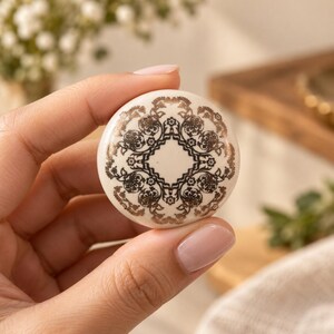 Puede incluir: Un pomo de cerámica redondo con una base de color crema y un patrón floral simétrico detallado impreso en un acabado de bronce metálico. El diseño consta de motivos botánicos complejos y repetitivos dispuestos en un diseño circular.