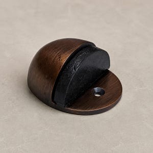 May include: A semi-circular door stop with a brushed bronze finish and a black rubber bumper insert. The metal base features a single pre-drilled screw hole for mounting, showcasing a smooth, curved dome shape designed to protect walls.