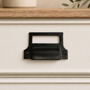 May include: A matte black metal bin pull handle with an integrated label holder frame. The hardware features a rectangular top section for a label and a curved grip area below, secured to a light-colored drawer front with two visible screws.