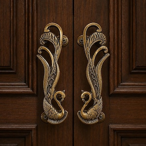 May include: A pair of antique brass door handles shaped like peacocks with intricate feather detailing and curved necks. The handles are mounted vertically on a dark brown wooden door with visible screws at the top and bottom of each piece.